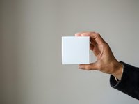 Adult male hand holding blank white square card against neutral background for branding, marketing, and promotional mockup purposes.