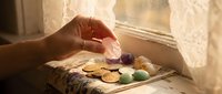 Hand placing colorful Easter eggs on vintage wooden tray by sunny window with warm natural light creating cozy spring holiday atmosphere for home decor.