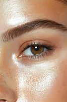 Close-up of glowing skin with natural freckles and hazel eye showing dewy, luminous complexion and well-groomed eyebrow for beauty and skincare advertising.
