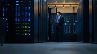 Professional African American male IT administrator working with tablet in modern server room with glowing network equipment and data center infrastructure.