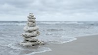 Balanced stone cairn tower on sandy beach with ocean waves and cloudy sky background symbolizing zen meditation mindfulness and spiritual harmony.