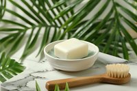 Natural soap bar in ceramic dish with bamboo toothbrush on marble surface surrounded by tropical palm leaves for eco-friendly bathroom decor.