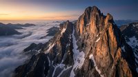 Dramatic mountain peak rising above sea of clouds at golden hour sunrise with rocky cliffs and snow patches creating breathtaking alpine landscape scenery.