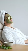 Young woman in white robe relaxing with green clay face mask and cucumber slices on eyes during spa skincare treatment.