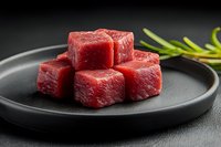 Fresh raw beef cubes on black ceramic plate with rosemary sprig. Close up view of marbled meat chunks ready for cooking. Dark moody background.