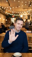 Middle-aged Caucasian businessman in navy sweater waving hello in cozy cafe with string lights overhead for greeting and hospitality concepts.