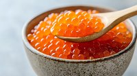 Fresh red caviar in ceramic bowl with wooden spoon, bright orange salmon roe pearls close up on light background, luxury seafood delicacy.