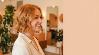 Happy young Caucasian businesswoman with short blonde bob haircut smiling in modern office environment with plants and warm lighting for corporate content.