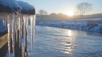 Glowing icicles hanging over frozen river at sunset, winter landscape with sunlight reflecting on ice surface and snowy riverbank in cold weather.