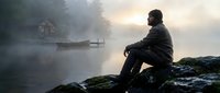 Man sitting on rocky shore contemplating misty lake at dawn with wooden boat in background creating peaceful solitude atmosphere for wellness and mindfulness concepts.