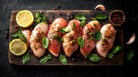 Raw chicken breast fillets seasoned with herbs, spices, lemon and fresh basil leaves arranged on rustic wooden cutting board against dark background.