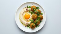 Creamy hummus topped with fried egg and crispy falafel balls garnished with fresh herbs on white plate for Middle Eastern cuisine and healthy eating.