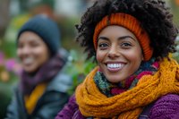 Young African American woman with curly hair wearing orange knit hat and colorful scarf smiling outdoors in autumn. Happy diverse people in warm winter clothing.