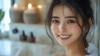 Young Asian woman with natural makeup and messy updo hairstyle smiling warmly at camera in soft window light, wellness spa atmosphere with candles and essential oils in background.