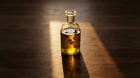 Glass bottle filled with golden whiskey or bourbon spirit, casting warm amber light on wooden surface. Suitable for beverage, alcohol, and liquor industry marketing.