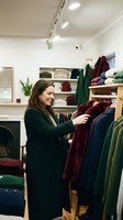 Woman shopping for clothing in modern retail store, browsing burgundy sweater on wooden rack with organized shelves of folded garments and accessories in background.