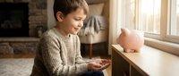 Young boy with light skin tone and dark hair, wearing a beige knit sweater, smiling while holding coins near a pink piggy bank on a windowsill. Soft natural light streams through windows in a cozy home interior. Concept for savings, financial education, and childhood money management.