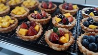 Fresh fruit mini tarts with crispy pastry shells filled with vanilla custard and topped with seasonal berries, peaches, and mangoes on cooling rack. Perfect for dessert buffet.