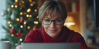 Middle-aged woman with short blonde hair and glasses wearing burgundy sweater, working on laptop surrounded by glowing holiday decorations and Christmas tree lights.