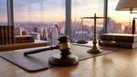 Wooden gavel and scales of justice on desk in modern law office with city skyline view during sunset. Legal consultation and attorney services concept.