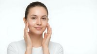 Young Caucasian woman applying facial cream with gentle hands touching cheeks in skincare routine on white background for beauty and wellness concepts.