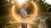 Young Caucasian man using smartphone on rural path during golden hour sunset with warm sunlight creating natural lens flare effect through trees.