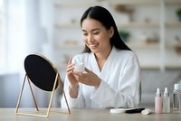 Young Asian woman in white bathrobe applying skincare cream while looking in mirror at vanity table with beauty products for morning routine.