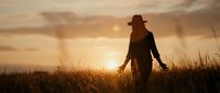 Silhouette of woman in hat walking through tall grass field at golden sunset with dramatic sky and warm lighting creating peaceful countryside atmosphere.