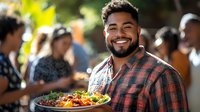 Young Hispanic man with beard smiling while holding colorful fresh salad plate at outdoor gathering with people in background enjoying social event.