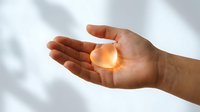 Human hands cupped together holding glowing orange light orb against soft white background for spiritual healing energy concepts.
