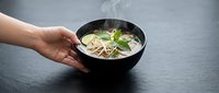 Hand holding black bowl of Asian noodle soup with bean sprouts and herbs on dark surface for culinary presentation and food styling concepts.