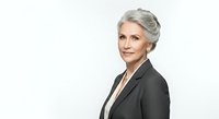 Mature woman with gray hair wearing dark blazer and white shirt, professional business portrait on white background.