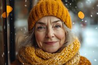 Senior woman with gray hair wearing mustard yellow knit beanie and scarf in snowy winter setting with bokeh lights, outdoor portrait.