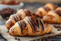 Fresh baked croissants drizzled with dark chocolate sauce on rustic burlap background, close-up view showing flaky pastry layers and glossy chocolate garnish.