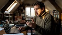 Man with dark hair and beard, wearing glasses and a brown knit sweater, gesturing while video conferencing on laptop in a cozy attic home office surrounded by bookshelves and natural light.