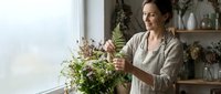 Middle-aged Caucasian woman arranging fresh flowers in bright kitchen interior. Natural light streaming through window creates warm atmosphere for home decor.