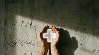 Close-up of hands forming a cross or plus symbol against weathered concrete wall with natural light and shadow, representing faith, healthcare, medical support, charity, or hope.