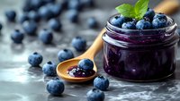 Fresh blueberry jam in glass jar with wooden spoon and scattered berries on rustic table. Homemade preserve with mint garnish for breakfast and cooking.