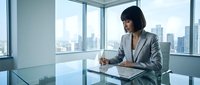 Professional Asian businesswoman in gray suit writing documents at modern glass conference table in corporate office with city skyline view through windows.