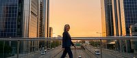 Young professional woman in dark business attire walking across elevated urban bridge at golden hour sunset, surrounded by modern skyscrapers and city traffic below.