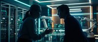 Two male technicians in dark uniforms examine a digital tablet while standing in a server room illuminated by neon teal lighting, surrounded by computer equipment and network infrastructure.