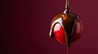 Chocolate syrup dripping over fresh red cherry creating glossy coating against burgundy background for dessert and confectionery advertising.