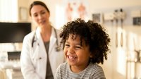 Young African American child with curly hair smiling during pediatric checkup with female doctor in medical office background.