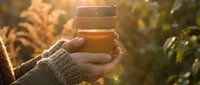 Hands in cozy knit sweater holding reusable coffee cup outdoors during golden hour sunset with warm autumn light filtering through foliage.