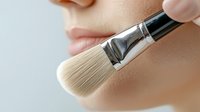 Close-up view of makeup brush applying foundation or powder on young woman's face, showing soft natural bristles and silver metal handle against smooth skin.