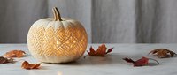 Carved white pumpkin with intricate geometric pattern sits on wooden surface with autumn leaves against rustic gray wood background for fall decor.