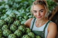 Young Caucasian female farmer in overalls smiling while holding fresh artichokes at local market. Green organic produce and sustainable agriculture concept.
