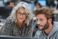 Senior woman with glasses and gray hair collaborating with young bearded man in office environment, looking at computer screen during professional discussion.