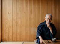 Senior man with gray hair and beard, African American ethnicity, wearing dark navy button-up shirt, seated at wooden table with contemplative expression, hand near chin, against warm wood paneling backdrop for business, leadership, wisdom, and professional portraits.
