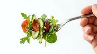 Fresh mixed green salad with cherry tomatoes and cucumber on fork held by hand against white background for healthy eating concepts.